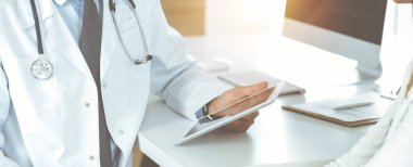 Doktor ve hasta, güneşli klinikteki masada otururken tıbbi muayene sonuçlarını tartışıyorlar. Yakın plan. İlaç geçmişini doldurmak için tablet bilgisayar kullanan bir erkek doktor..