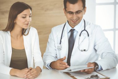 Doktor ve hasta klinikteki masada otururken tıbbi muayene sonuçlarını tartışıyorlar. Genç bir kadının ilaç geçmişini doldurmak için tablet bilgisayar kullanan bir erkek doktor. Tıptaki veriler