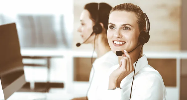 Mujer rubia representante de servicio al cliente y su colega están consultando a los clientes en línea utilizando auriculares. Centro de llamadas y concepto de gente de negocios — Foto de Stock