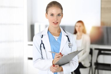 Kadın - doktor, arka plandaki güneşli klinikte tıbbi geçmiş kayıtlarını doldurmak için pano kullanıyor.