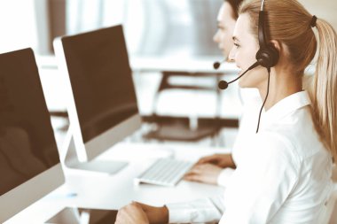 Sarışın kadın müşteri hizmetleri temsilcisi ve meslektaşı internetteki müşterilerine kulaklık kullanarak danışıyorlar. Çağrı merkezi ve iş adamları kavramı
