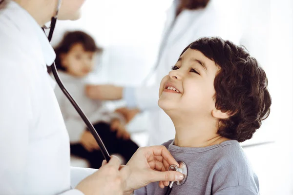 Médico-mujer examinando a un niño paciente por estetoscopio. Lindo chico árabe y su hermano en la cita con el médico. Concepto de medicina —  Fotos de Stock