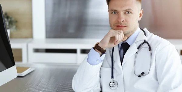 Arzt sitzt am Arbeitsplatz in sonniger Klinik hinter einem Computer — Stockfoto