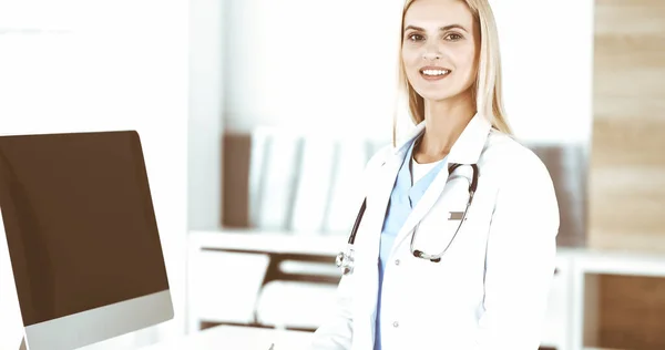 Blond female physician is standing at her workplace near desktop computer. Woman-doctor is excited and happy of her profession. Medicine concept — Stock Photo, Image