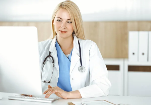 Mulher-médico no trabalho enquanto sentado na mesa no hospital ou na clínica. Médico alegre loira usando computador desktop — Fotografia de Stock