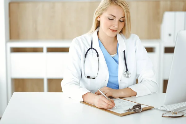 Retrato de mulher-médico no trabalho enquanto sentado na mesa na clínica. Médico alegre loiro preenchendo formulário médico ou prescrição — Fotografia de Stock