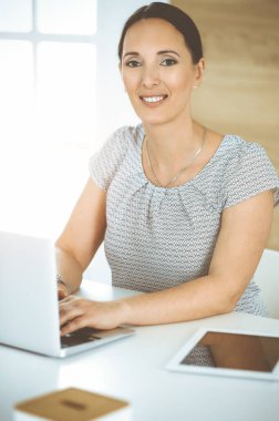 Neşeli gülümseyen iş kadını güneşli ofisteki masada otururken bilgisayarla çalışıyor. Orta yaşlı kadın avukat ya da denetçi.