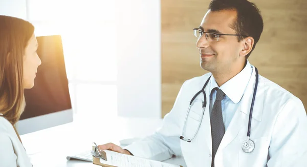 Médecin et patient discutant des résultats de l'examen médical assis au bureau dans une clinique ensoleillée — Photo