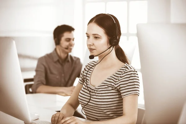 Casual vestido jovem mulher usando fone de ouvido e computador enquanto conversa com clientes on-line. Grupo de operadores a trabalhar. Call center, conceito de negócio — Fotografia de Stock