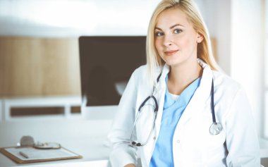 Güneşli klinikte masa başında oturan kadın doktor. Sarışın, neşeli doktor yardıma hazır.