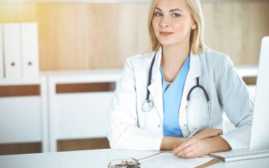 Güneşli klinikte masa başında oturan kadın doktor. Sarışın, neşeli doktor yardıma hazır.