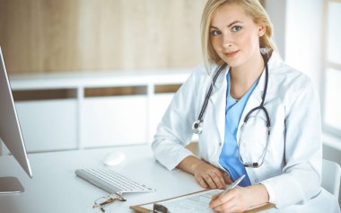 Güneşli klinikte masa başında oturan kadın doktor. Sarışın, neşeli doktor ilaç geçmişi formunu dolduruyor.