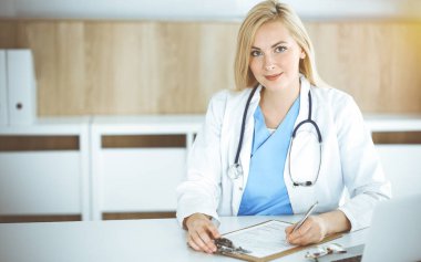 Güneşli klinikte masa başında oturan kadın doktor. Sarışın, neşeli doktor ilaç geçmişi formunu dolduruyor.