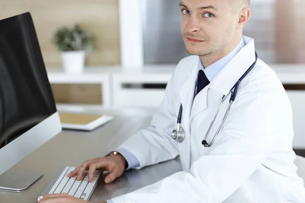 Dokter man zit achter een computer op het werk in zonnige kliniek. — Stockfoto