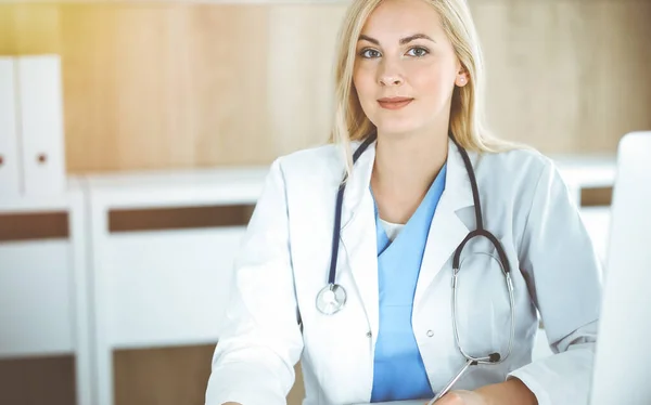 Mulher-médico no trabalho enquanto sentado na mesa na clínica ensolarada. Médico alegre loiro pronto para ajudar — Fotografia de Stock
