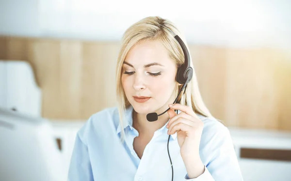 Blonde business woman sitting and communicated by headset in call center in sunny office. Concept of telesales business