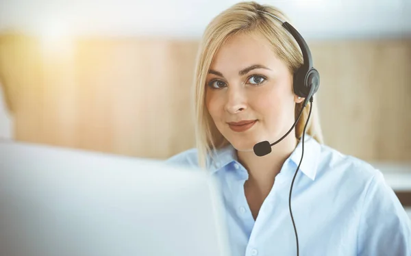 Blonde business woman sitting and communicated by headset in call center in sunny office. Concept of telesales business