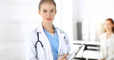 Kadın - doktor, arka plandaki güneşli klinikte tıbbi geçmiş kayıtlarını doldurmak için pano kullanıyor.