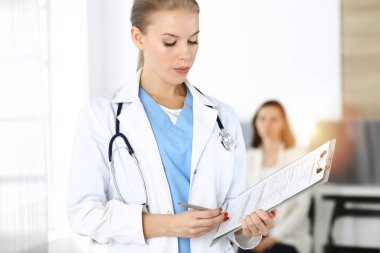 Kadın - doktor, arka plandaki güneşli klinikte tıbbi geçmiş kayıtlarını doldurmak için pano kullanıyor.