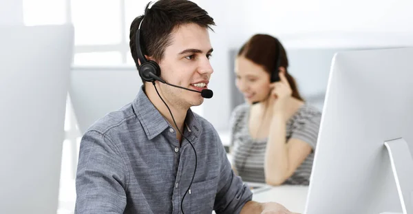 Hombre joven vestido casual usando auriculares y computadora mientras habla con los clientes en línea. Grupo de operadores en el trabajo. Centro de llamadas —  Fotos de Stock