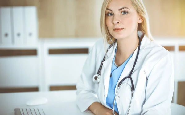 Mujer-médico en el trabajo mientras está sentado en el escritorio en la clínica soleada. Rubia alegre médico llenar la historia de la medicación formulario de registro — Foto de Stock