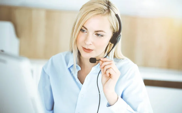 Blonde business woman sitting and communicated by headset in call center in sunny office. Concept of telesales business