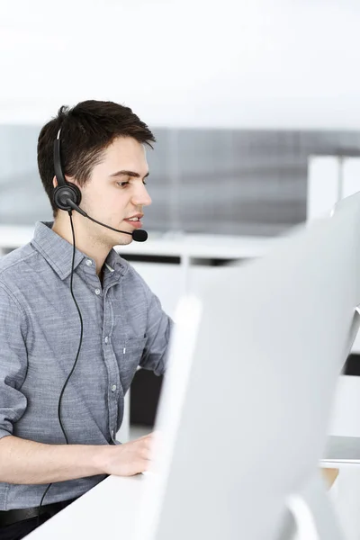 Hombre joven vestido casual usando auriculares y computadora mientras habla con los clientes en línea. Call center, concepto de negocio —  Fotos de Stock