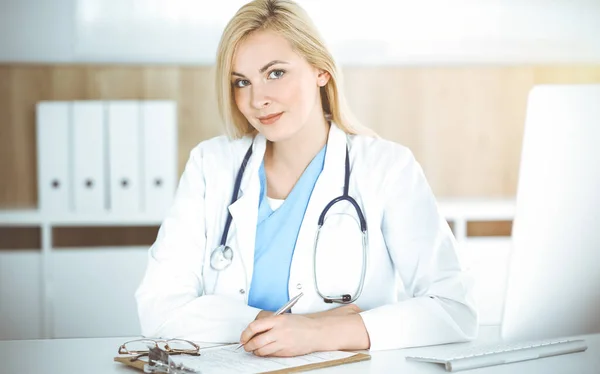 Mujer-médico en el trabajo mientras está sentado en el escritorio en la clínica soleada. Rubia médico alegre listo para ayudar — Foto de Stock