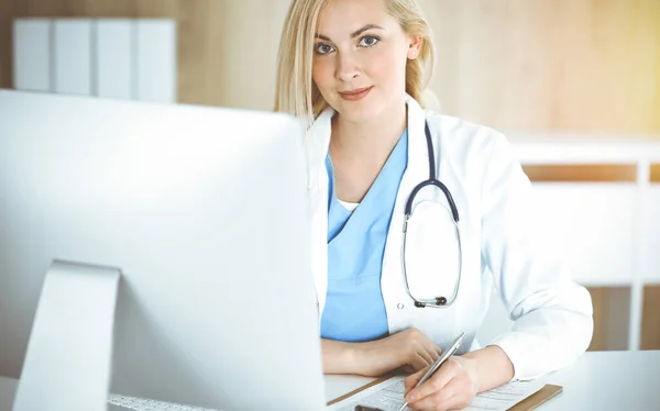 Žena-doktor v práci, zatímco sedí za stolem na slunné klinice. Blondýny veselý lékař vyplňování léku historie záznam formě — Stock fotografie