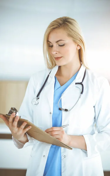 Femme-médecin debout dans une clinique ensoleillée et remplissant le formulaire d'historique de médicaments. Données en médecine — Photo