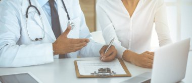 Doktor ve hasta, güneşli klinikteki masada otururken tıbbi muayene sonuçlarını tartışıyorlar. Yakın plan. Erkek doktor pano kullanıyor.