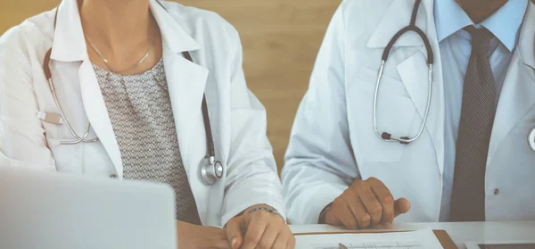 Два неизвестных врача обсуждают проблемы лечения, сидя за столом в солнечном больничном офисе, крупным планом — стоковое фото