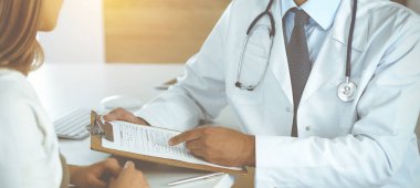 Doktor ve hasta, güneşli klinikteki masada otururken tıbbi muayene sonuçlarını tartışıyorlar. Yakın plan. Erkek doktor pano kullanıyor.