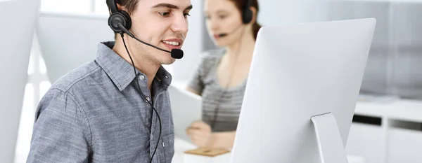 Hombre joven vestido casual usando auriculares y computadora mientras habla con los clientes en línea. Grupo de operadores en el trabajo. Centro de llamadas —  Fotos de Stock