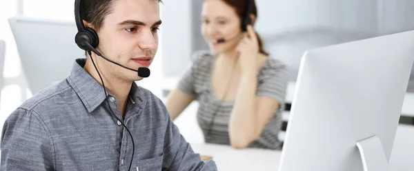 Hombre joven vestido casual usando auriculares y computadora mientras habla con los clientes en línea. Grupo de operadores en el trabajo. Centro de llamadas —  Fotos de Stock