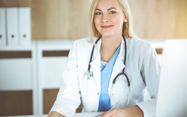 Kvinna-läkare på jobbet medan du sitter vid skrivbordet i solig klinik. Blond glad läkare redo att hjälpa — Stockfoto