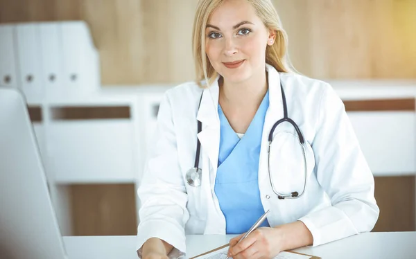 Mujer-médico en el trabajo mientras está sentado en el escritorio en la clínica soleada. Rubia alegre médico llenar la historia de la medicación formulario de registro — Foto de Stock