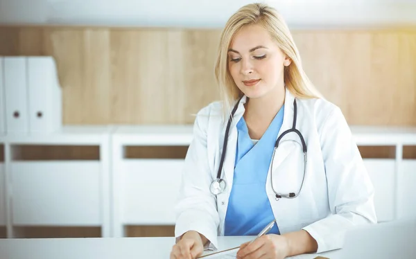 Mujer-médico en el trabajo mientras está sentado en el escritorio en la clínica soleada. Rubia alegre médico llenar la historia de la medicación formulario de registro — Foto de Stock