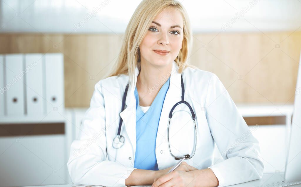 Woman-doctor at work while sitting at the desk in sunny clinic. Blonde cheerful physician ready to help