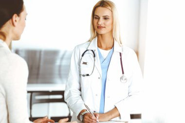 Sunny Hastanesi 'ndeki kadın doktor kadın hastaya danışmaktan mutluluk duyar. Sarışın doktor, pano kullanırken tıbbi geçmişi ve sınav sonuçlarını kontrol ediyor.