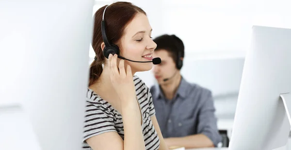 Casual dressed young woman using headset and computer while talking with customers online. Group of operators at work. Call center, business concept — Stock Photo, Image