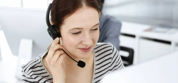Casual dressed young woman using headset and computer while talking with customers online. Group of operators at work. Call center, business concept — Stock Photo, Image