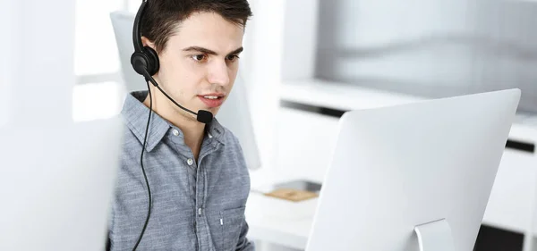 Casual ντυμένος νεαρός άνδρας χρησιμοποιώντας ακουστικά και υπολογιστή, ενώ μιλάμε με τους πελάτες σε απευθείας σύνδεση. Τηλεφωνικό κέντρο, επιχειρηματική ιδέα — Φωτογραφία Αρχείου