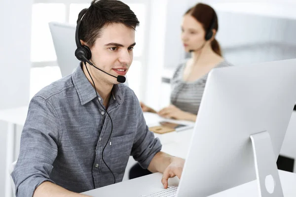 Hombre joven vestido casual usando auriculares y computadora mientras habla con los clientes en línea. Grupo de operadores en el trabajo. Centro de llamadas —  Fotos de Stock