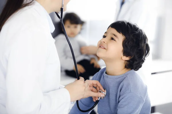 Médico-mujer examinando a un niño paciente por estetoscopio. Lindo chico árabe y su hermano en la cita con el médico. Concepto de medicina — Foto de Stock