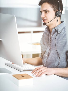 Güneşli ofiste müşterilerle konuşurken kulaklık ve bilgisayar kullanan sıradan giyinmiş genç bir adam. Çağrı merkezi, iş konsepti