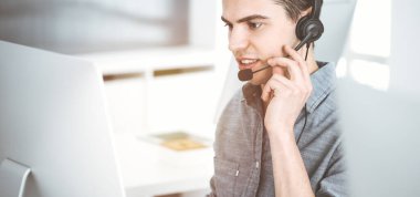 Güneşli ofiste müşterilerle konuşurken kulaklık ve bilgisayar kullanan sıradan giyinmiş genç bir adam. Çağrı merkezi, iş konsepti