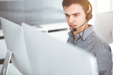 Güneşli ofiste müşterilerle konuşurken kulaklık ve bilgisayar kullanan sıradan giyinmiş genç bir adam. Çağrı merkezi, iş konsepti