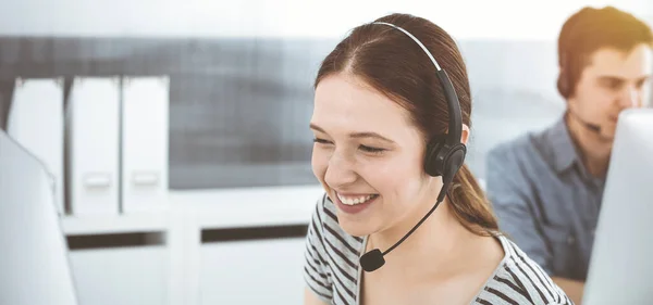 Casual dressed young woman using headset and computer while talking with customers online. Group of operators at work in sunny office. Call center