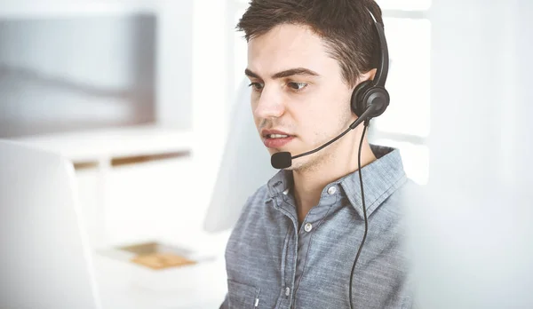 在阳光明媚的办公室里，穿着便服的年轻人一边在网上与顾客交谈，一边用耳机和电脑。呼叫中心，商业概念 — 图库照片